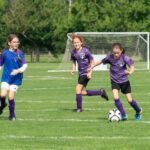Pier to Pier Bike Ride Young soccer competitors in purple and blue uniforms on a green field. White goal posts are visible in the background near a natural green tree line.