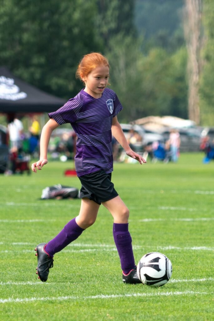 A young soccer player wearing a purple uniform kicks the ball on a grassy field, with a casual outdoor setting and blurred greenery in the Pacific Northwest background.