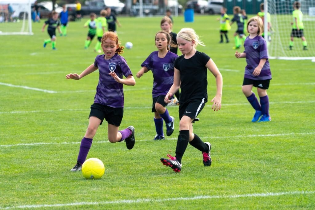 A yellow soccer ball on the field with players from two teams competing. Young athletes in purple and black uniforms focus on the game.