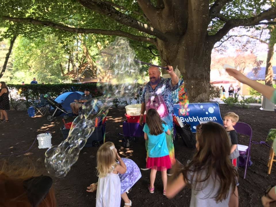 A group of children watch a performer creating bubbles in a sunny outdoor park. A sign reading 'Bubbleman' is visible near the audience.