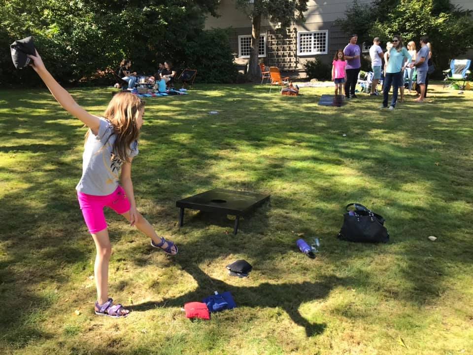 A person tossing a beanbag toward a cornhole game in a sunny backyard. Other people gather nearby on the grass with a house in the background.