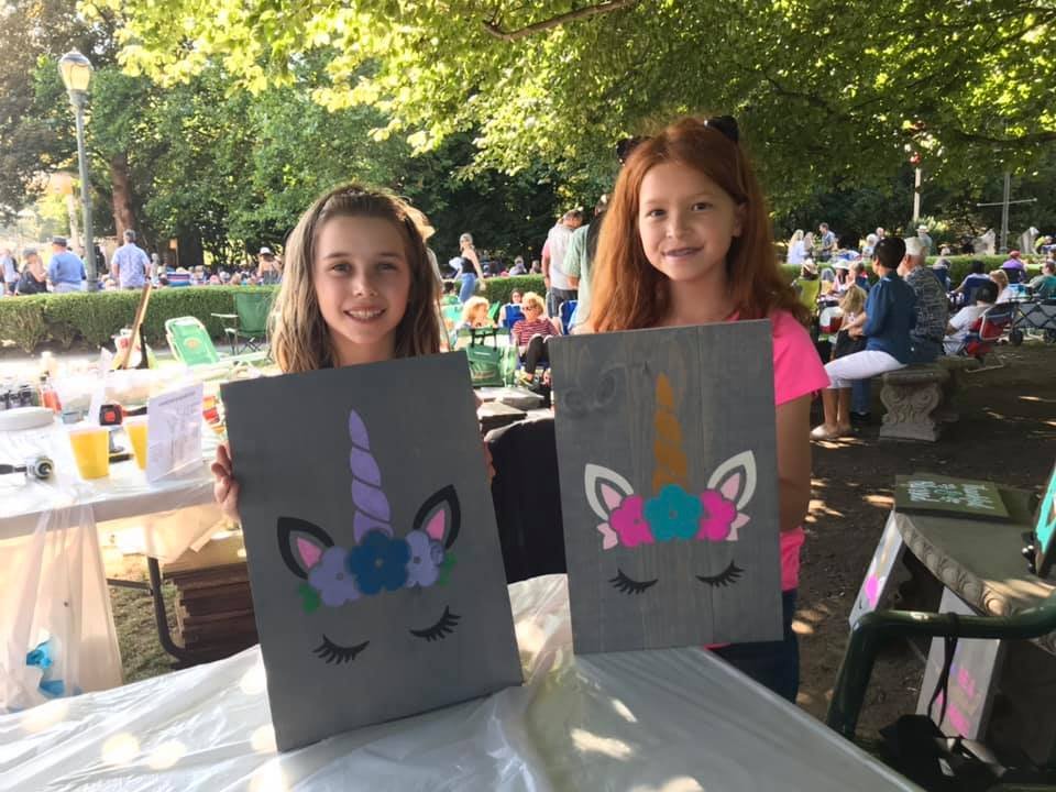 Two girls proudly display handmade paper unicorn headboards at an outdoor community event.