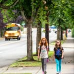 Two students walking down a wide sidewalk lined with mature trees, with a yellow school bus driving on the street behind them.