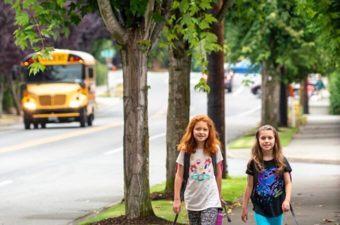 Two students walking down a wide sidewalk lined with mature trees, with a yellow school bus driving on the street behind them.