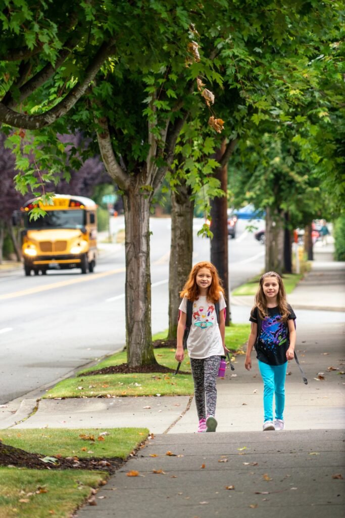 Two students walking down a wide sidewalk lined with mature trees, with a yellow school bus driving on the street behind them.