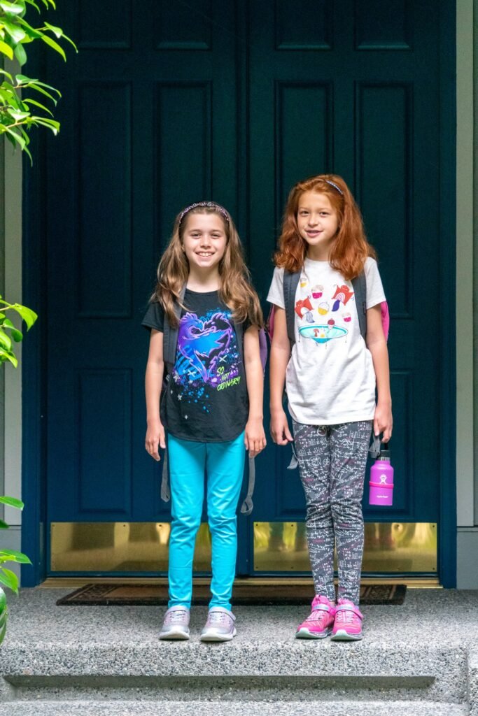 Two children standing in front of a blue door wearing backpacks and colorful clothing.