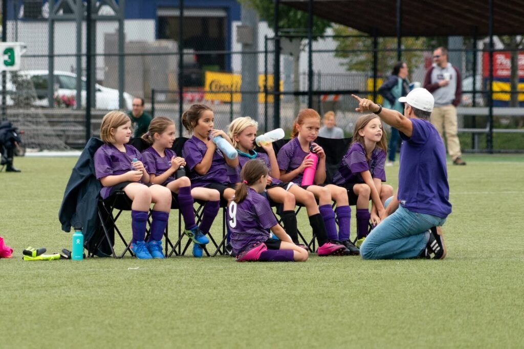 A youth soccer team in purple uniforms listens to a coach on the sideline.