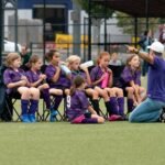 A youth soccer team in purple uniforms listens to a coach on the sideline.