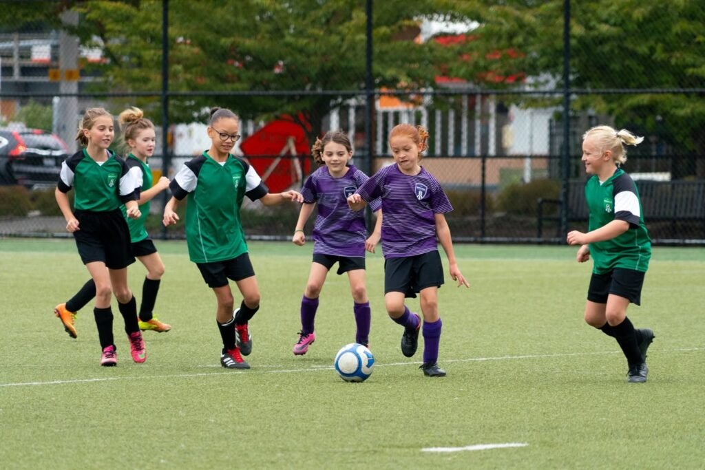 Young soccer players compete on a grassy field, with teams in green and purple jerseys chasing the ball.
