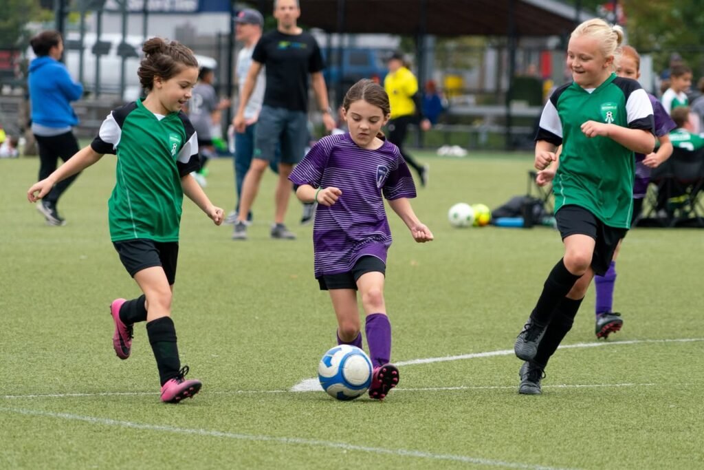 Children in green and purple uniforms playing soccer on a grassy field, with one player kicking a ball.
