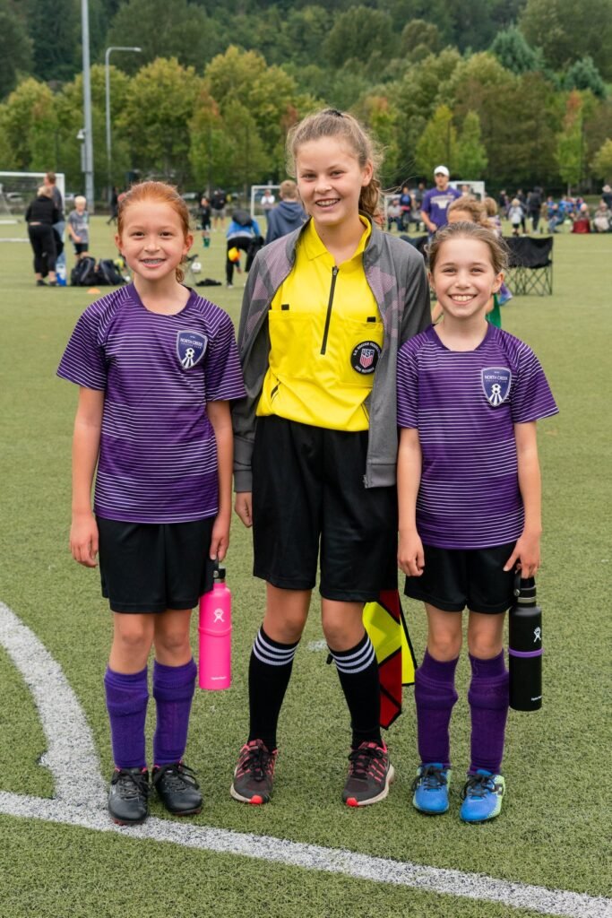 A group photo of a soccer coach and two young players in purple and yellow uniforms on a grass field.