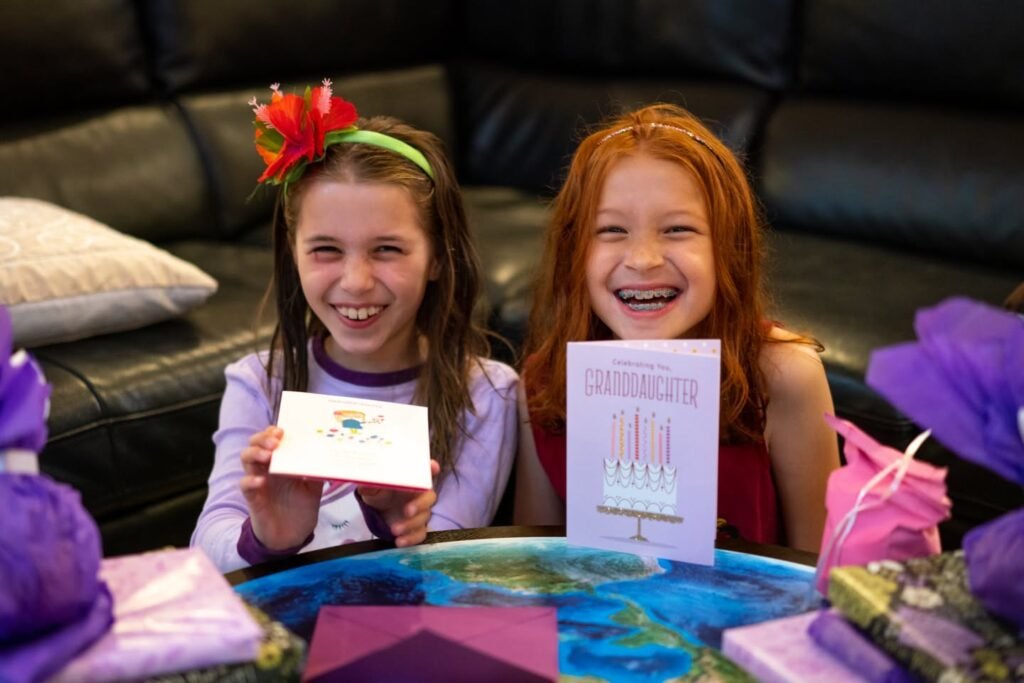 Two children smile while holding handmade birthday cards, sitting at a table with purple decorations in a cozy living room.
