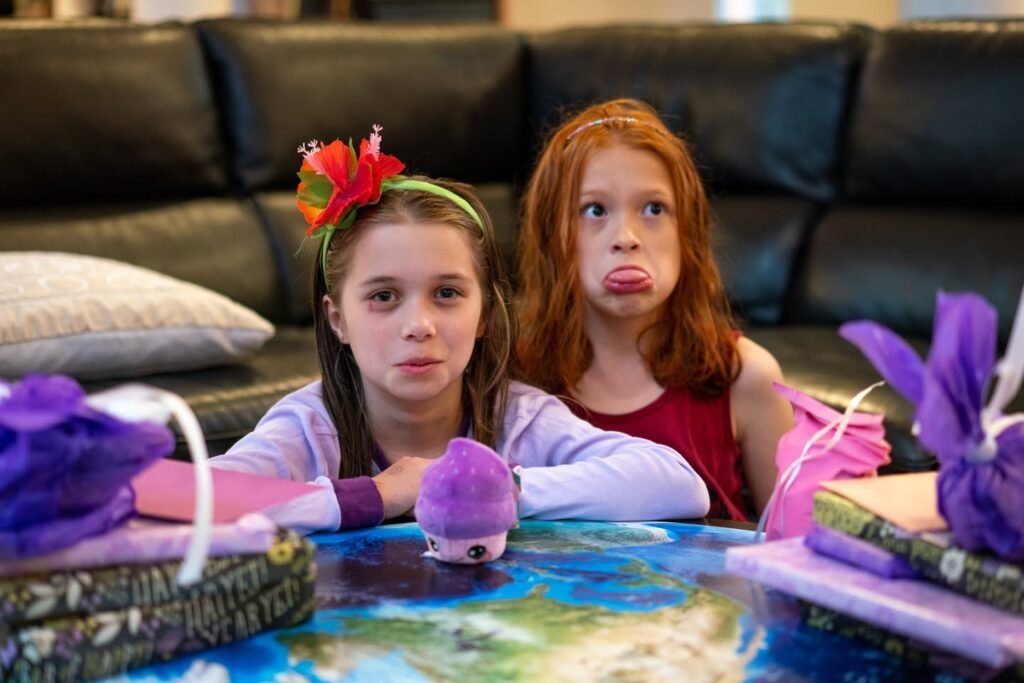 Two young girls making faces together at a table with a world map and decorated gifts during a family gathering.