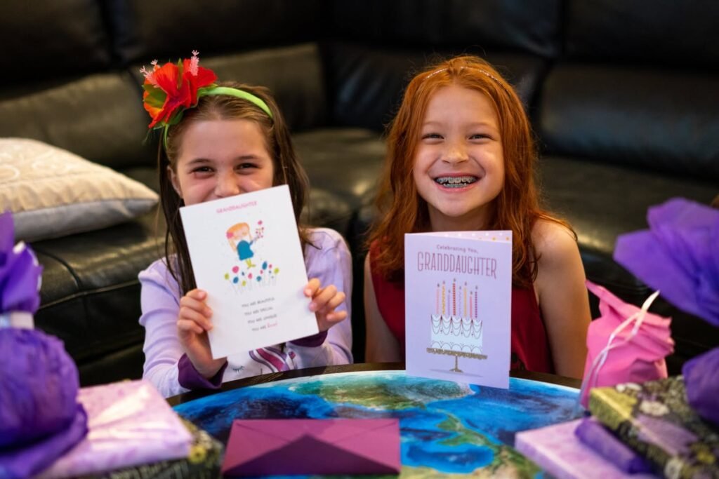 Two young people smiling with birthday cards and purple balloons by a black leather couch.