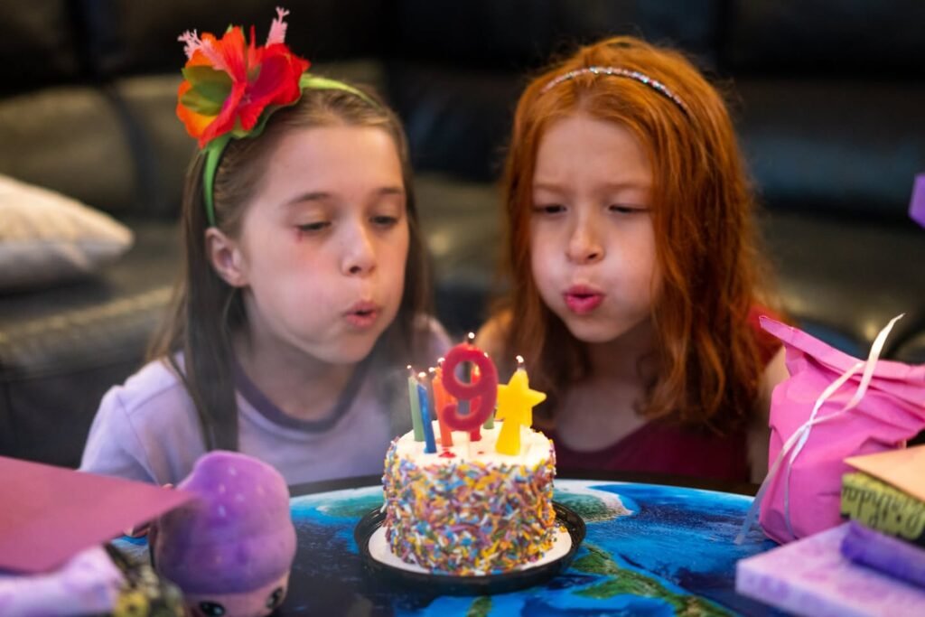 Two young sisters wear floral headbands and look down at a small birthday cake.