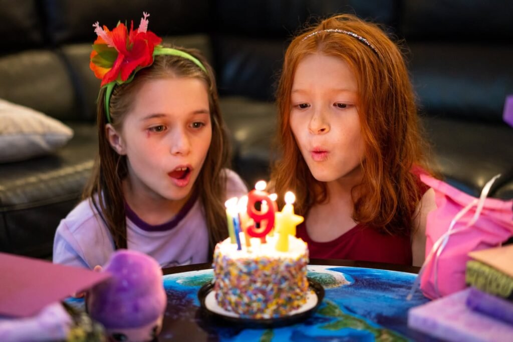 Two children lean over a glittery birthday cake with lit candles shaped like the number 9. A wrapped gift and a headband with a red flower are visible.