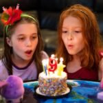 Two children lean over a glittery birthday cake with lit candles shaped like the number 9. A wrapped gift and a headband with a red flower are visible.