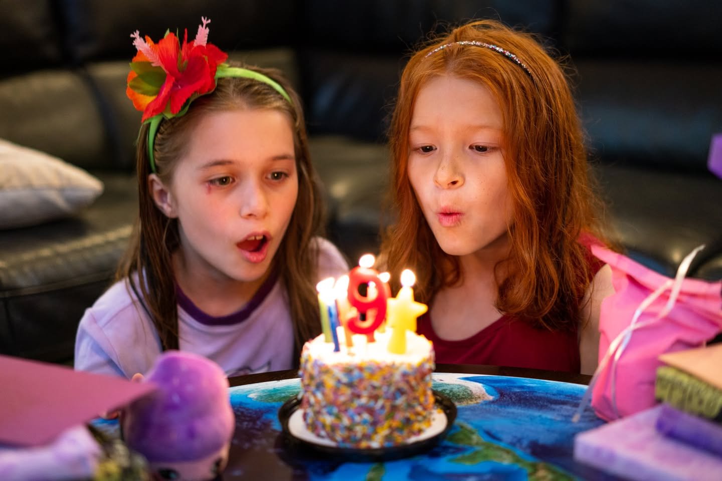 Nine Two children lean over a glittery birthday cake with lit candles shaped like the number 9. A wrapped gift and a headband with a red flower are visible.
