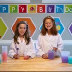 Children in lab coats conduct colorful experiments at a Happy Birthday display.