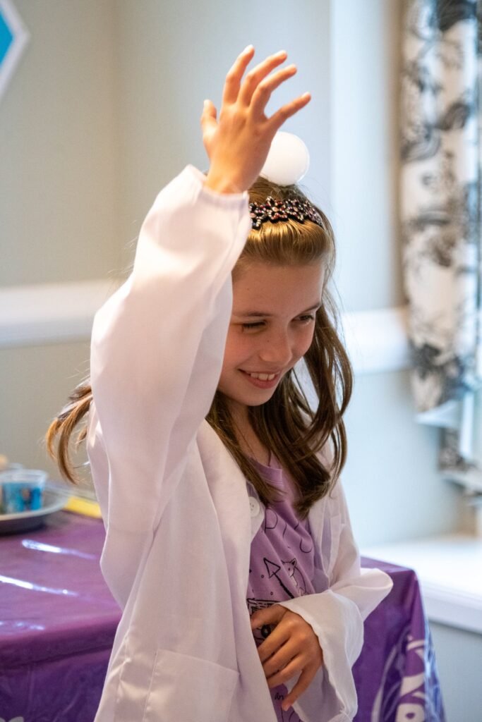 A smiling child in a white princess costume and tiara raises a hand playfully at a party.