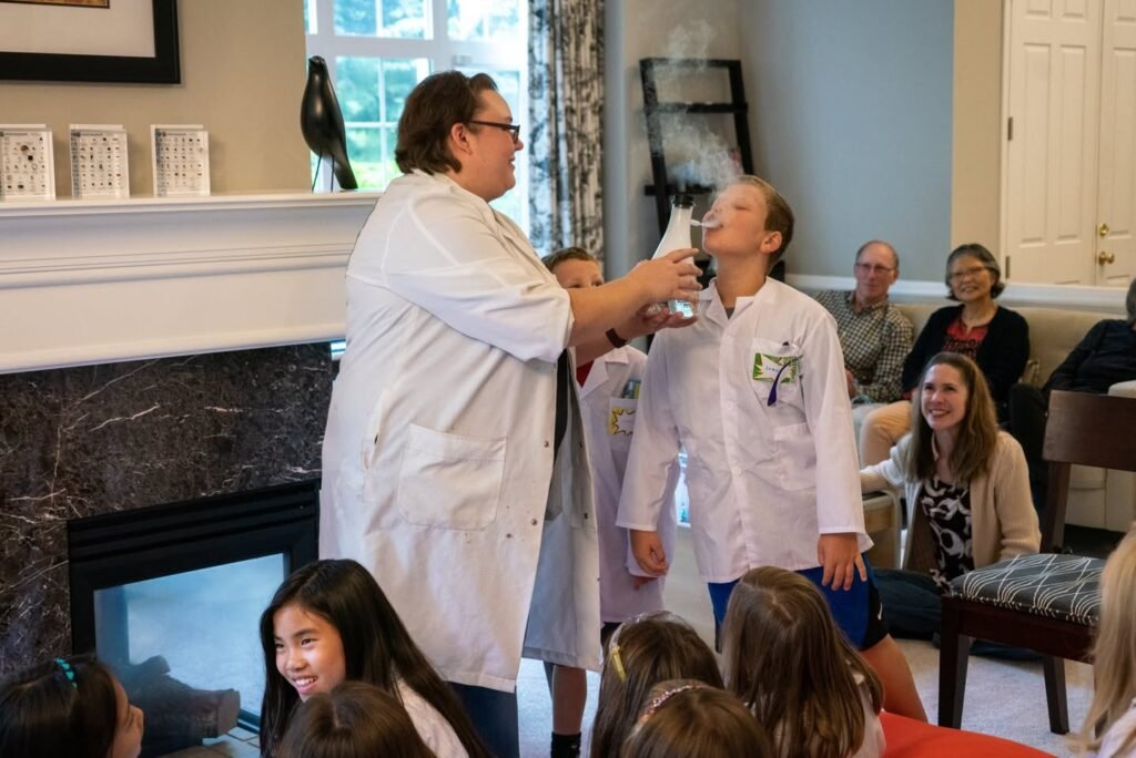 A person in a lab coat demonstrates a smoke experiment to children while adults observe from a sofa.