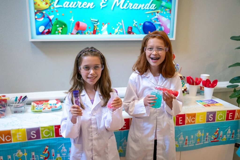 Two children in white lab coats holding experimental supplies at a table decorated with colorful 'Science' banners. A teal sign behind them reads 'Lauren & Miranda' above illustrations of science equipment.