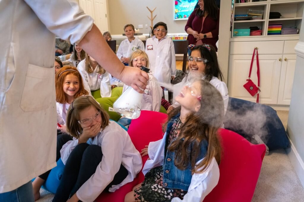 A group of children in lab coats participate in a science experiment with foggy dry ice clouds at Red Chairs during an educational event.