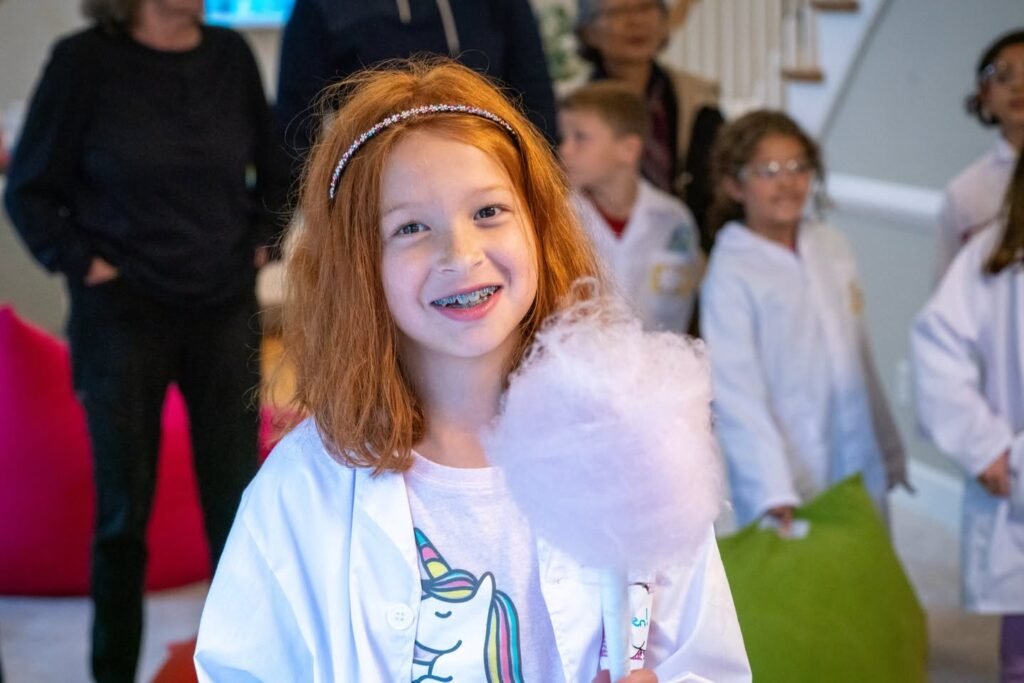 A young student wearing a white lab coat smiles while holding a prop. Other students in lab coats stand nearby at an event.