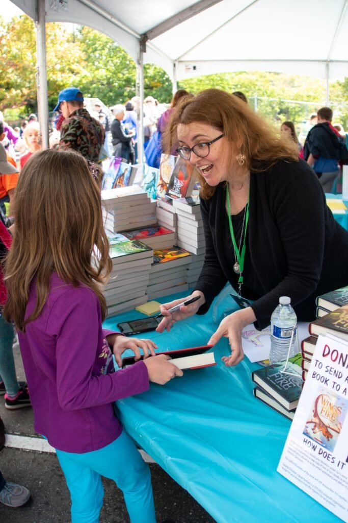 A smiling author in a black sweater offers books to a young reader at an outdoor book fair table. The scene is set under a white canopy with stacks of books visible.