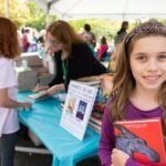 Pusheen’s Birthday A smiling girl in a purple cardigan holding a red book at an outdoor book fair table.