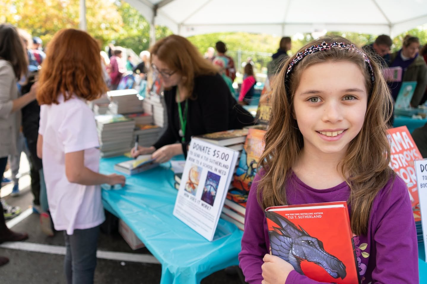 Seattle Children’s Book Festival A smiling girl in a purple cardigan holding a red book at an outdoor book fair table.