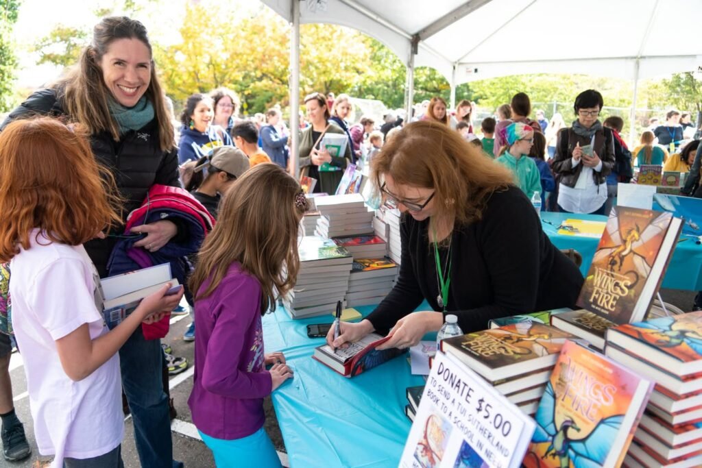 A woman signs a book for children under a white tent at a crowded outdoor fair. Stacks of 'World of Thieves' books are visible. A smiling woman stands in the crowd.