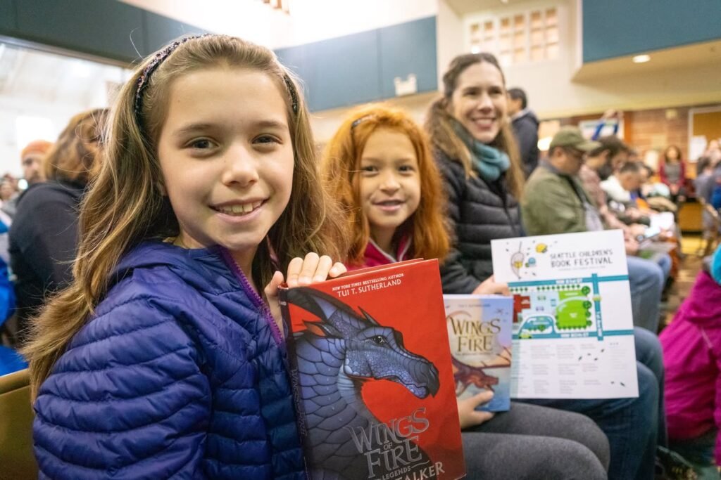 A smiling young person holds a Wingfire book at a bookstore event.