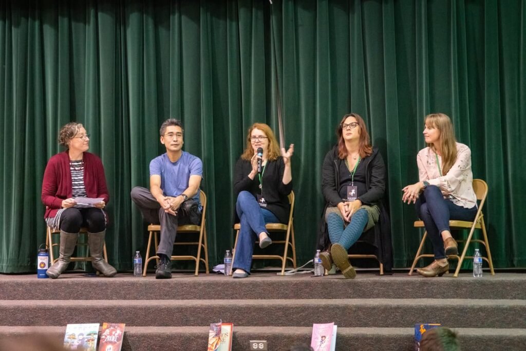 Five people seated on a stage against a green curtain for a panel discussion. Books are displayed on the steps in front of them.
