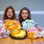 Two smiling girls sitting behind a table filled with colorful bakery-themed plush toys.
