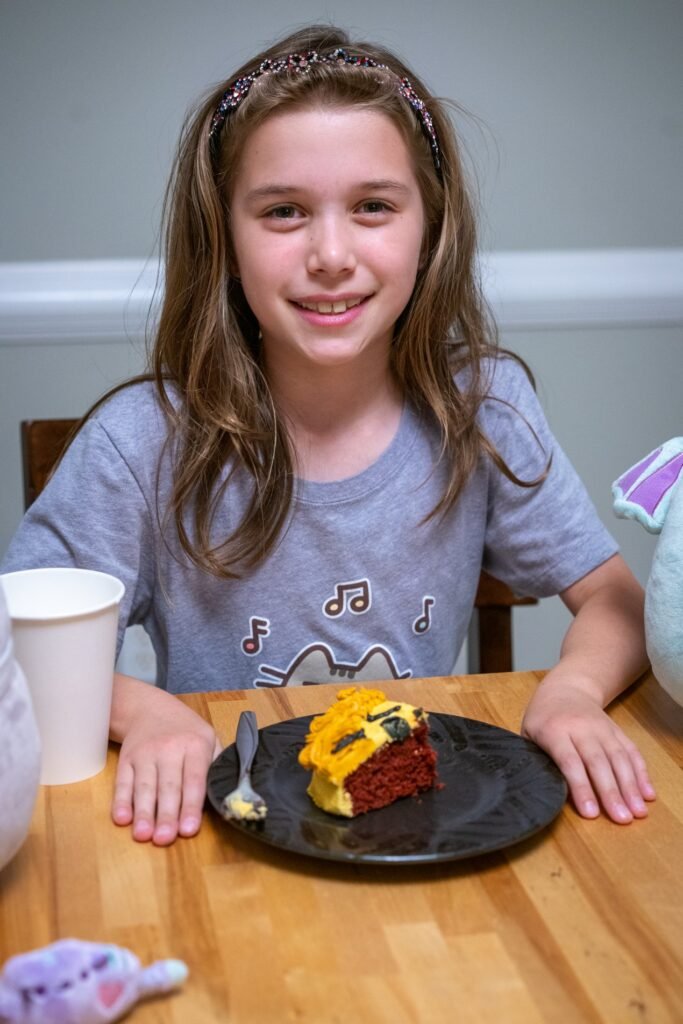 A young person smiles while sitting at a table with a slice of yellow cake, surrounded by cups and plush toys.
