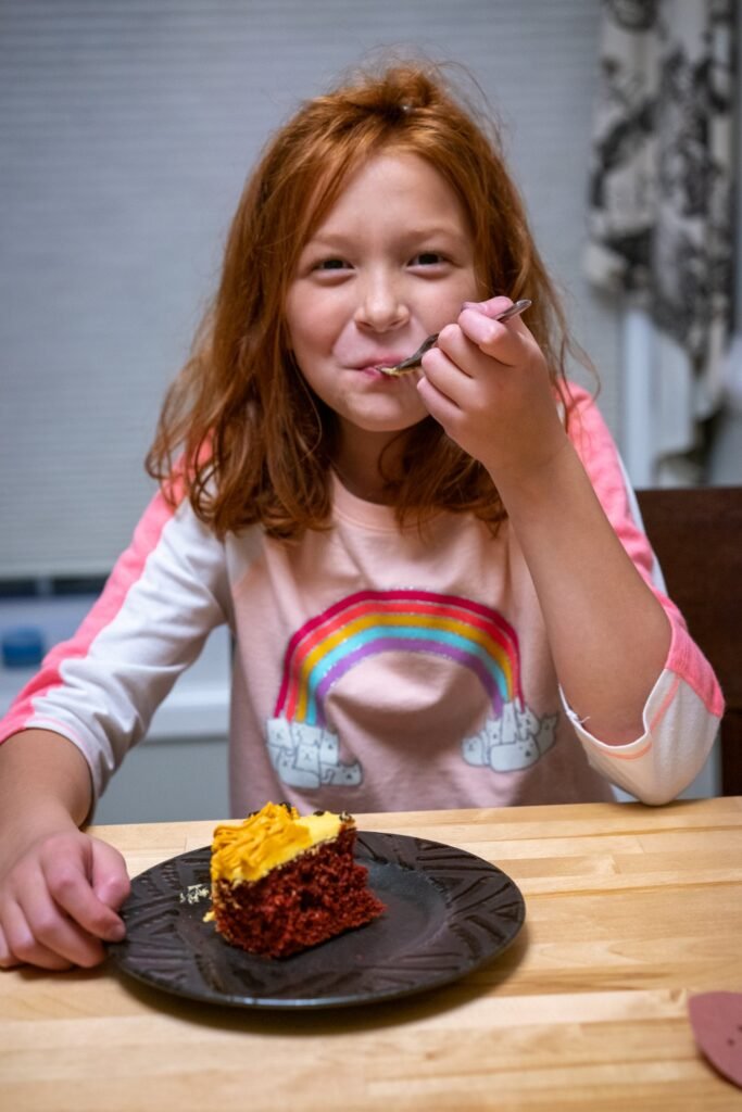 A young girl wearing a rainbow shirt enjoys a slice of cake at a wooden table, using a fork to take a bite.