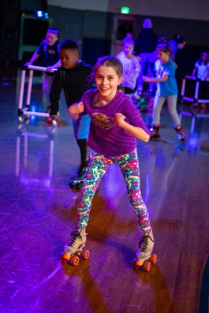 Children wearing colorful outfits and roller skates glide across an indoor rink illuminated by purple neon lights.