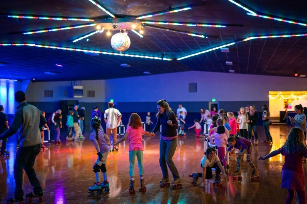 People skating on a roller rink with colorful overhead lighting and a classic disco ball. The polished wooden floor reflects the vibrant light patterns.