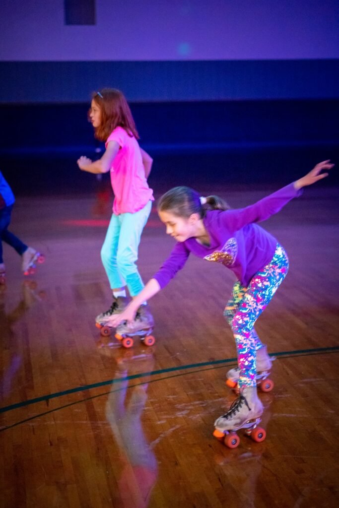 A child performs a skate move on a polished wooden floor behind another skater in a pink shirt.