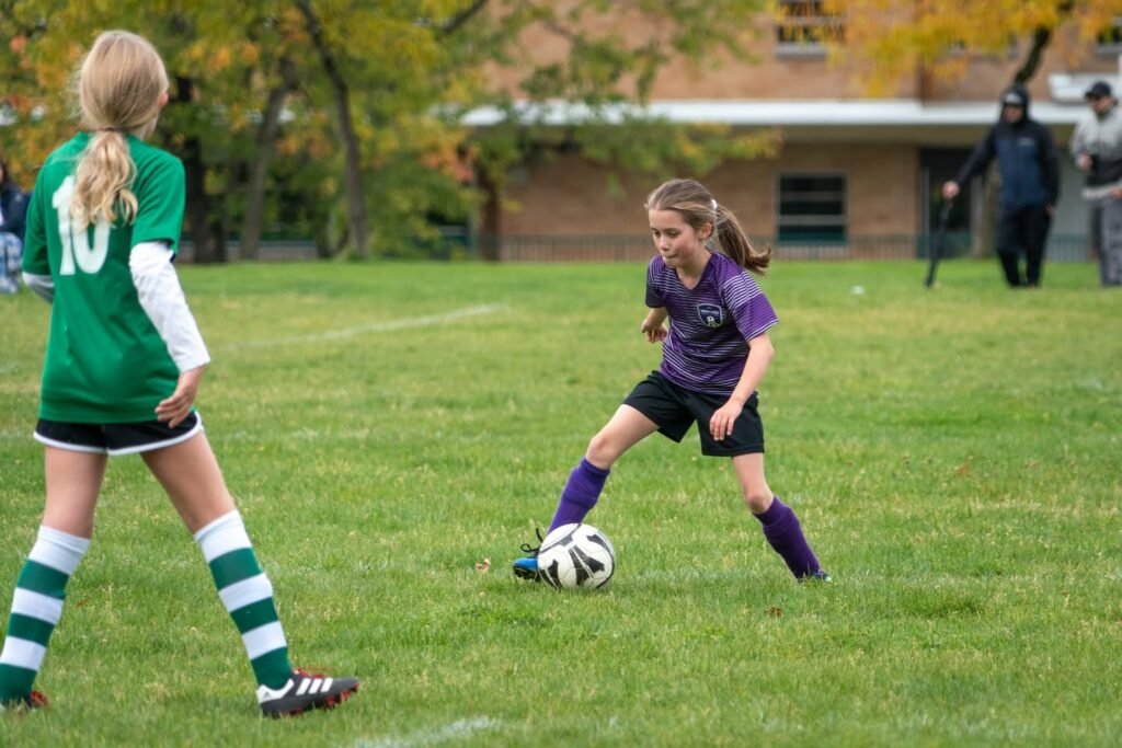 A close-up of a young soccer player dribbling a ball on a green field with an opponent in a green jersey nearby.