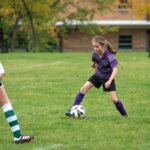 A close-up of a young soccer player dribbling a ball on a green field with an opponent in a green jersey nearby.