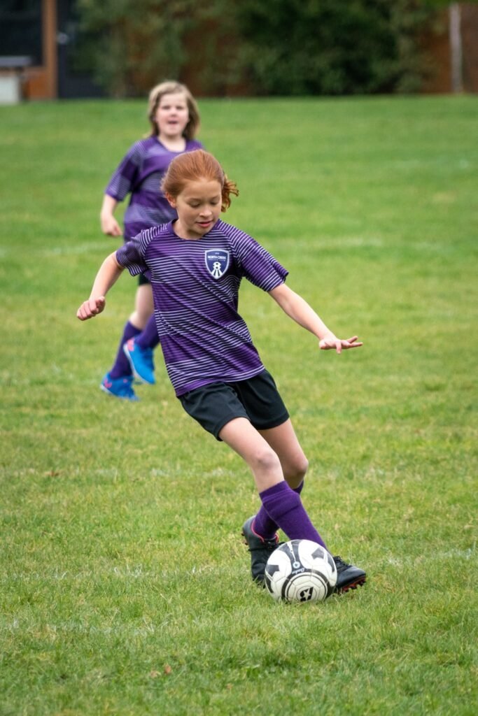 Young person in purple soccer uniform kicks a ball on a grassy field.
