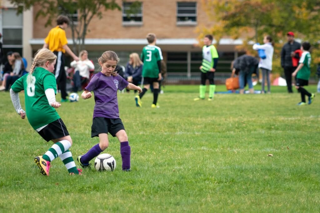 A young soccer player in a purple jersey competes with a player in a green jersey on a grassy field, with spectators in the background.
