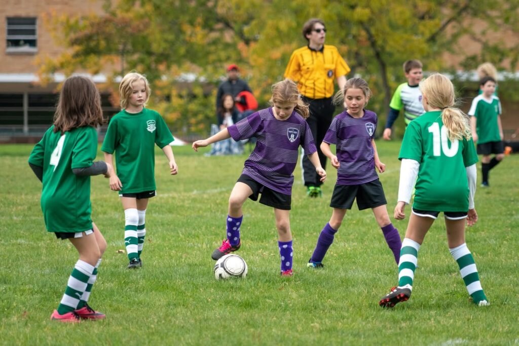 Young players in green and purple uniforms compete on a grass field during a soccer game. A referee in yellow observes from the background near autumn trees.