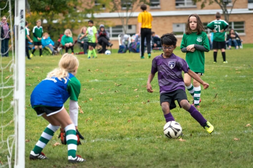 A young player in purple dribbles the ball towards the goal. The goalkeeper is ready on the field during a youth soccer match.