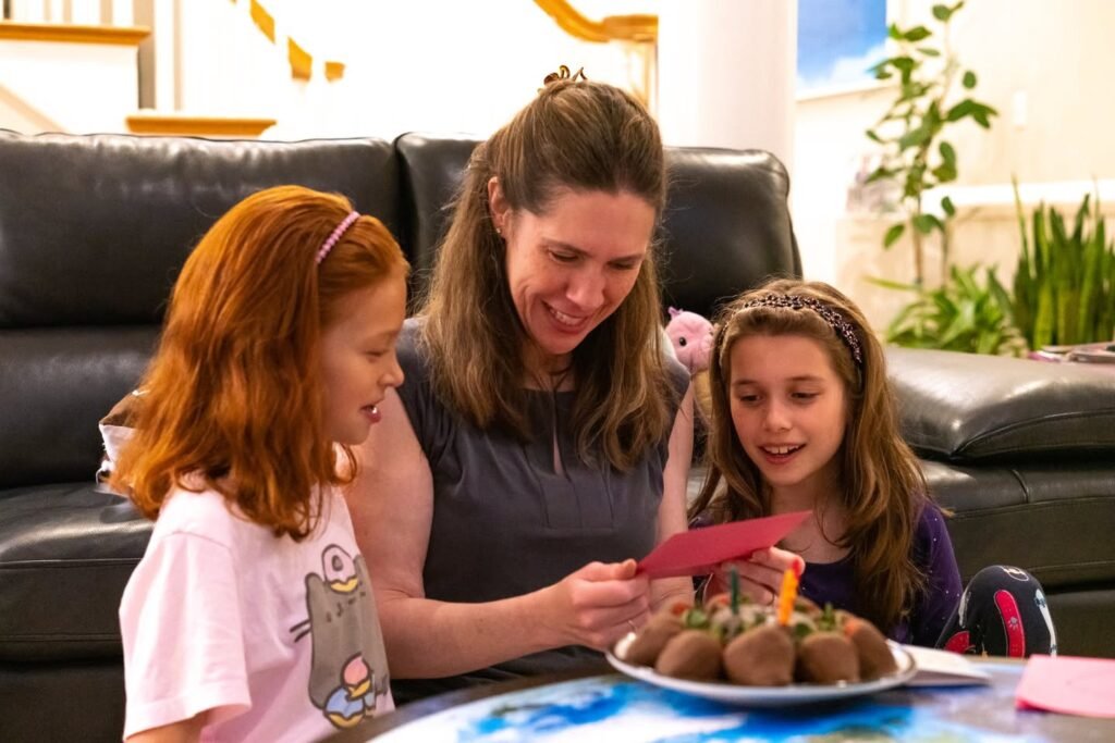 A woman and two young girls are enjoying a celebratory moment with a plate of chocolate truffles on a table. The setting features a black leather couch and homey decor.
