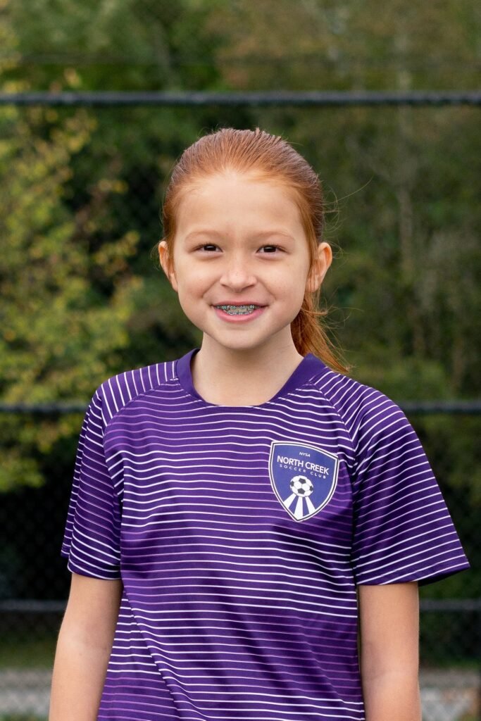 A smiling young athlete wearing a purple striped jersey with a rhythm gym logo. The background features a blurred outdoor setting with a fence.