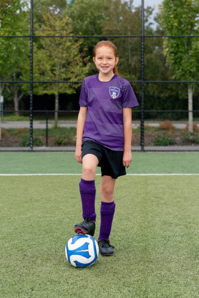 A young soccer player in a purple uniform stands over a blue and white ball on an artificial turf field.