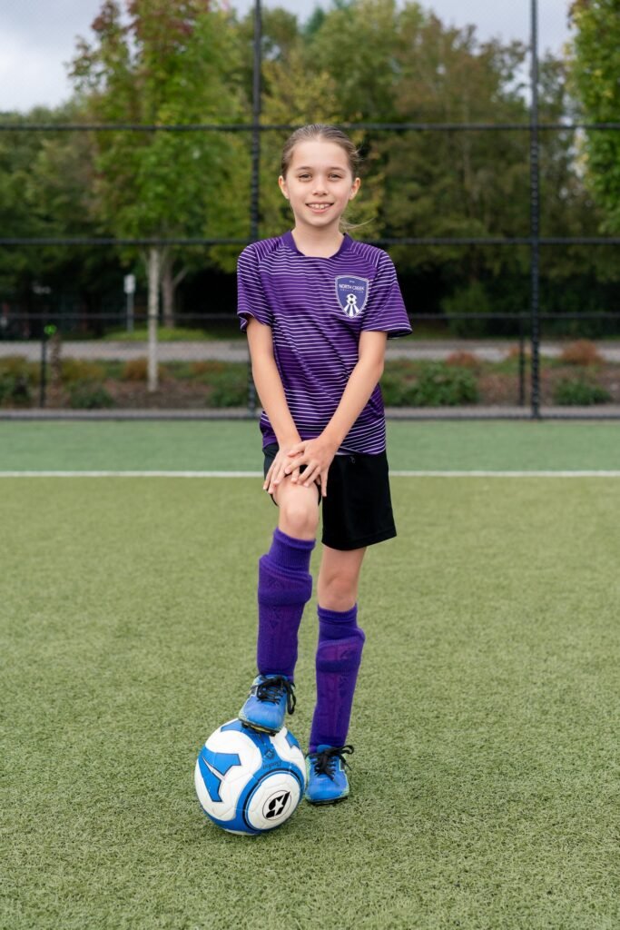 A young person in a purple soccer uniform poses on an artificial turf field with a blue and white ball.
